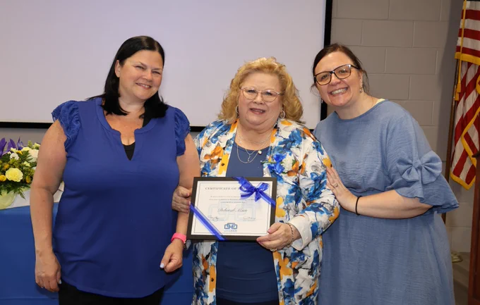 Three women standing together, one holding a framed certificate tied with a blue ribbon, with a table of flowers and an American flag in the background.
