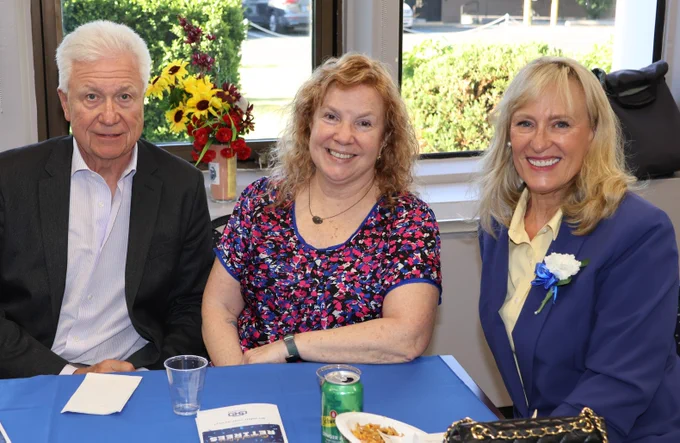 Three people sitting at a table with blue tablecloth, enjoying a casual gathering with snacks and drinks near a window.
