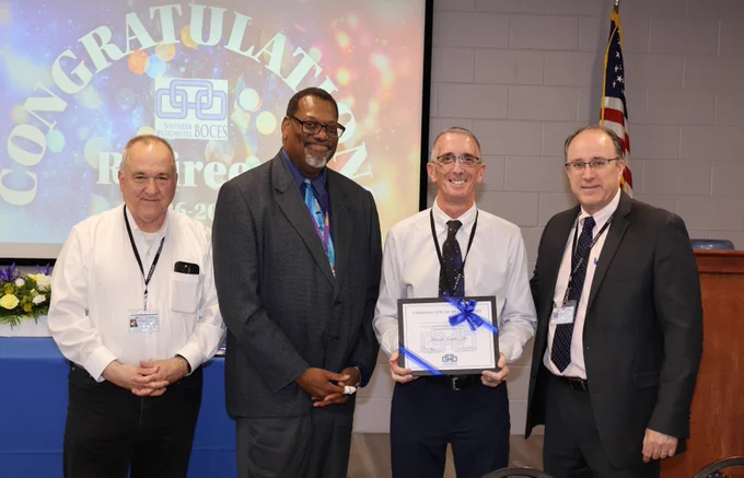 Four men in business attire stand together at an award ceremony, one holding a framed certificate with a blue ribbon, with a congratulatory message projected behind them.