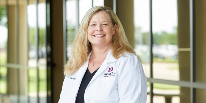 Dr. Michele Garant wears a white coat embroidered with Texas A&M Health Women's Care Plus logo while smiling at the camera