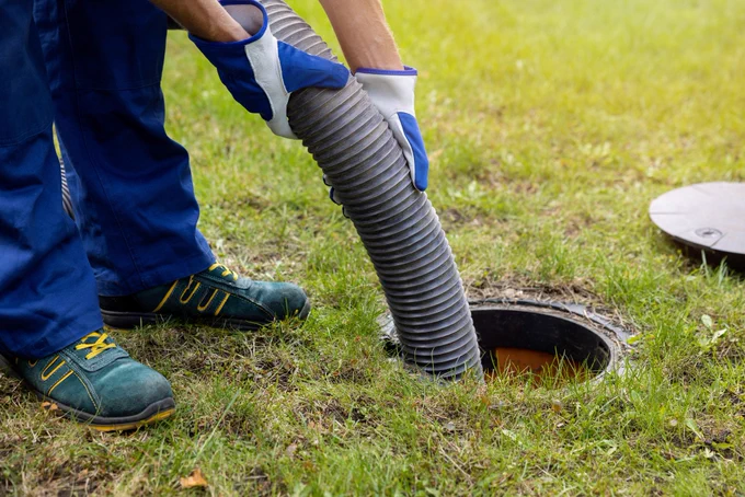 Worker wearing gloves inserts a flexible hose into an open septic tank on a grassy lawn.