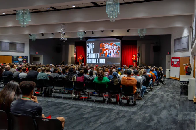 A large crowd of students seated in an auditorium facing a stage with two presenters and a large projector screen reading "2026 NEW STUDENT ORIENTATION".