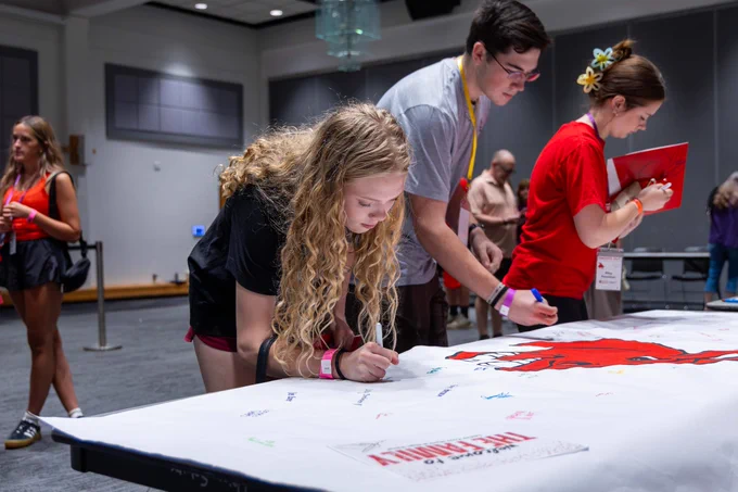Students leaning over a long table to sign a large banner featuring a red Mule logo illustration.