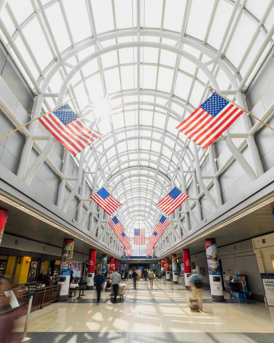 A spacious airport terminal with a high arched ceiling and numerous large United States flags hanging overhead. People are walking through the bright corridor, and information booths line the sides.