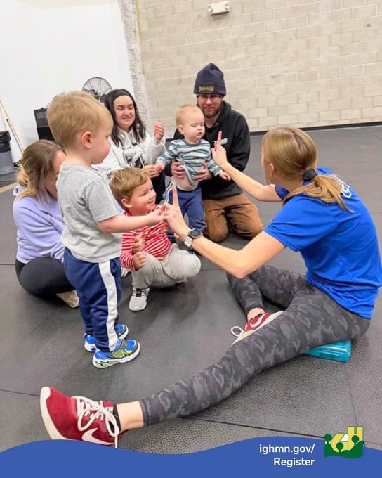 A woman in a blue shirt and camo leggings sits on the floor engaging with a group of young children and adults in a gym setting.