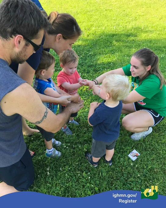 A group of adults and toddlers interacting and holding hands on a grassy field during an outdoor activity.