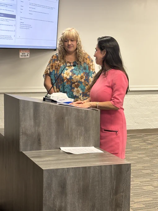 Woman at a podium speaks during a meeting while another woman stands beside her listening.