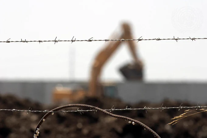 Barbed wire in the foreground with a blurred excavator behind it during construction of the separation wall in the occupied West Bank.
© UN Photo