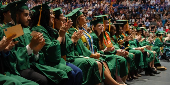 FCC graduates in green caps and gowns sit in rows, clapping during the Commencement ceremony with a large audience in the background.