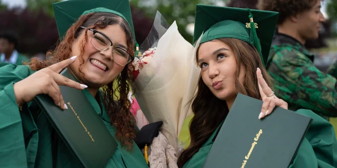 Two FCC graduates in green caps and gowns hold diplomas and a bouquet, posing with peace signs.