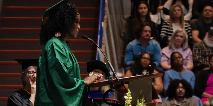 Graduate in green cap and gown delivering a speech at a podium decorated with yellow and white flowers during Commencement.