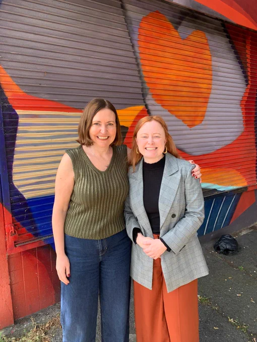 Vicky (left) and Nina (right), Director of UK Feminista, standing side by side in front of a colourful mural.