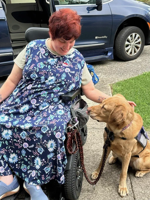 Dori- a white woman in a power wheelchair, pets her service dog after voting.