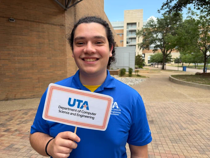 a man smiles while holding a sign with the UTA CSE logo on it