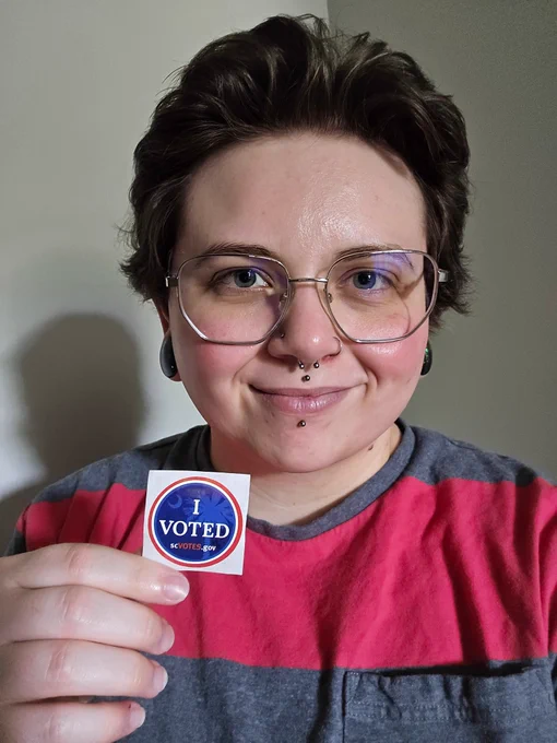 Jayden- a white person with glasses and facial piercings smiles with their I voted sticker.