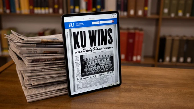 A stack of newspapers holds up an ipad featuring the online portal showing a historic student newspaper cover page. 