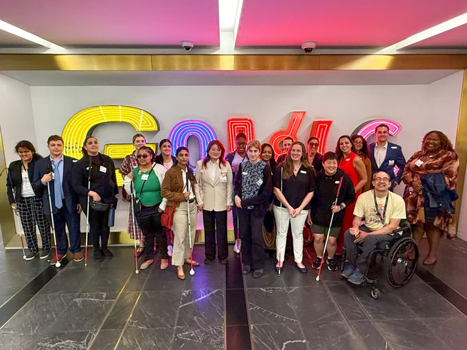 Group photo of 17 VISIONS participants and staff and three Google staff posing in front of a neon Google sign in the lobby of their office on 8th Avenue