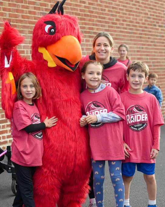 A large, red costumed mascot posing with a group of smiling children wearing matching pink shirts.