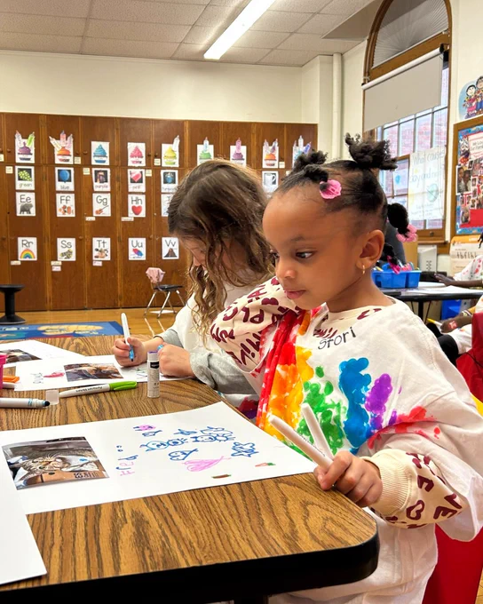 Young girl sitting at a desk in a classroom, working on an art project and wearing a colorful tie-dye shirt.