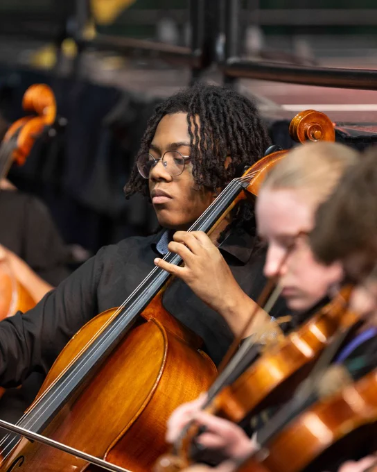 A student playing the cello on a stage, surrounded by other musicians playing instruments.