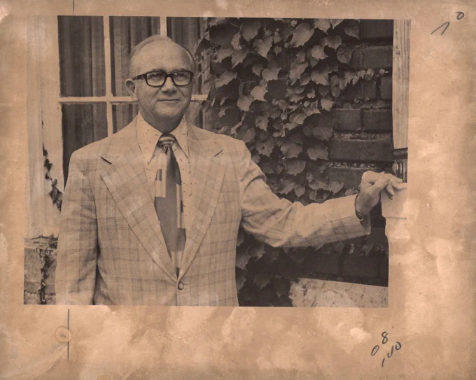 A yellowed photograph of Bob Ashton standing in a checkered sports jacket that is buttoned in the front. He is wearing a patterned collared shirt and tie under the jacket. Ashton is standing in front of a brick building with a window directly behind him. There are plants growing up the brick wall behind Ashton and he has his hand resting on part of the wall. He is wearing black glasses and is smiling.