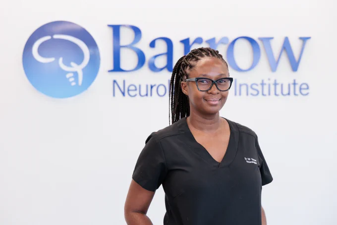 Dr. Sandane Chauke-Korea poses for a photo in front of the Barrow logo. She is wearing black scrubs embroidered with her name.