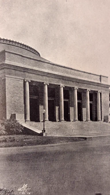 A 1926 photograph of Cornell's Bailey Hall, a neoclassical building with a columned entrance and wide steps. The structure is surrounded by manicured bushes and trees, with a clear plaza in the foreground.