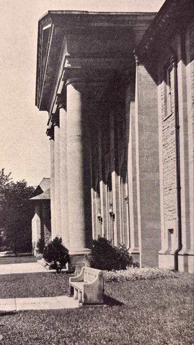 Side view of Cornell's Goldwin Smith Hall as seen in 1926, featuring tall classical columns and a stone bench on a grassy area in the foreground. Trees line the walkway beside the building.