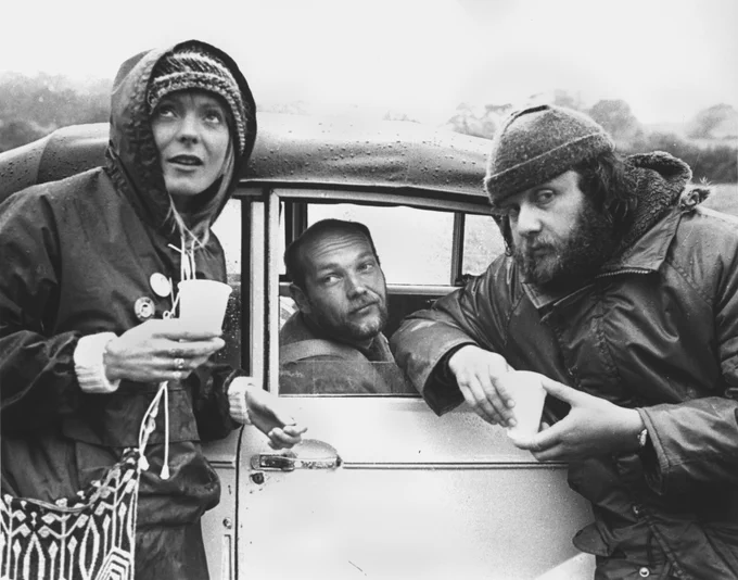 Three individuals are gathered around a vintage car. One person is seated inside, looking towards the camera, while the other two stand outside holding cups. They are dressed warmly, suggesting a chilly or rainy day. The setting appears to be outdoors, possibly in a rural area.