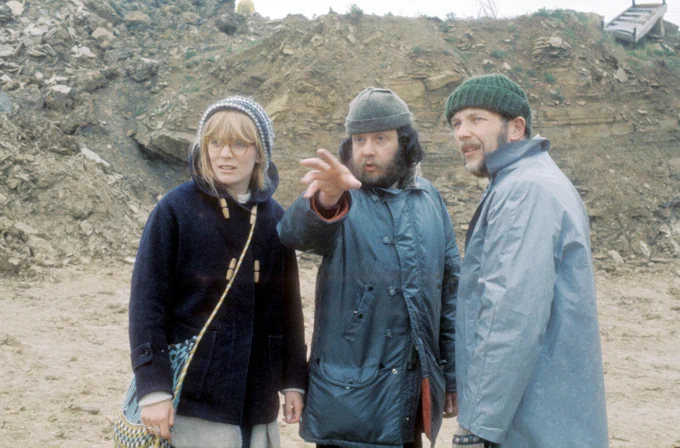 Three people stand on a rocky outdoor area. The person in the middle is gesturing into the distance whilst others look in the same direction. They are dressed in warm, casual clothing.