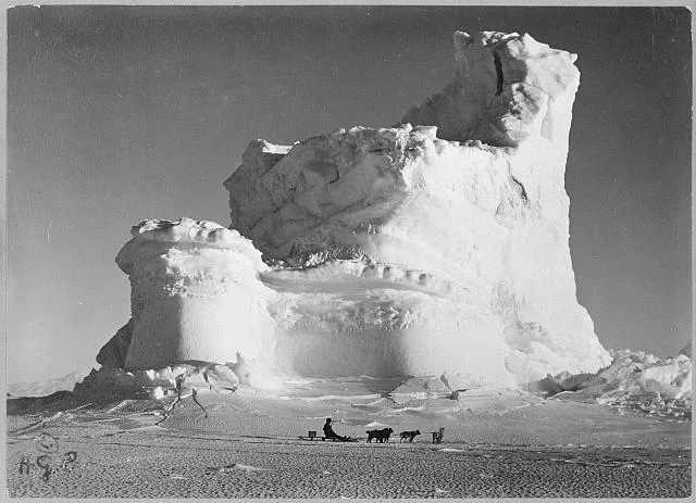 Photograph shows a man on a dogsled in the foreground and a castle-shaped mountain of ice in the background, during the British Antarctic Expedition.
