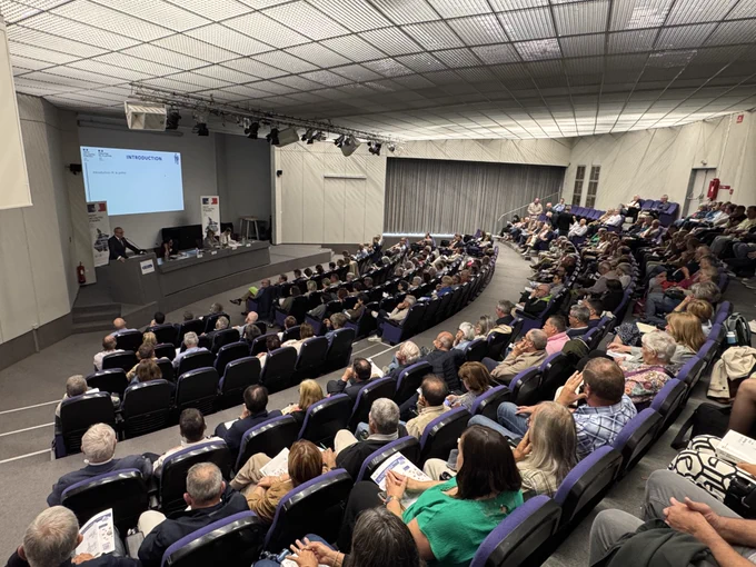 Salle de conférence remplie de plusieurs centaines d’élus locaux, assis dans un amphithéâtre lors d’une réunion d’information. Sur scène, le préfet des Hautes-Pyrénées, la procureure de la République, la secrétaire générale et la directrice de cabinet sont installés derrière une table face à un écran de projection affichant une présentation.