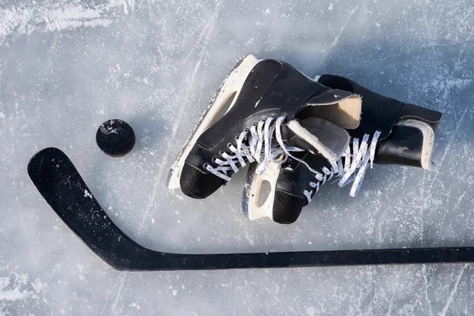 Close-up of black hockey skates, a hockey stick, and a puck resting on scratched ice.