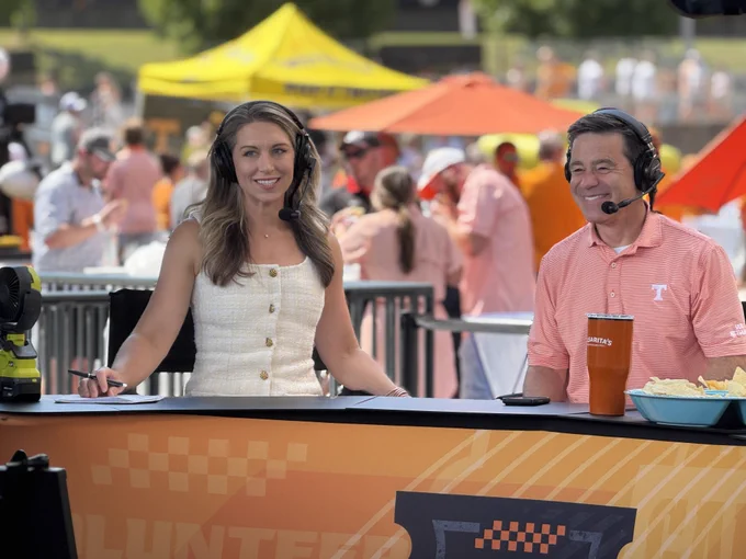 Two sports commentators sit at an outdoor desk, wearing headsets. They are smiling as they discuss, with a lively crowd and vibrant umbrellas behind them.