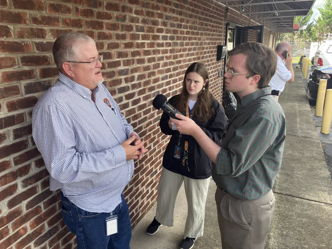 A man in a checkered shirt is interviewed by a young man with a microphone. A woman stands nearby, looking attentive against a brick wall backdrop.