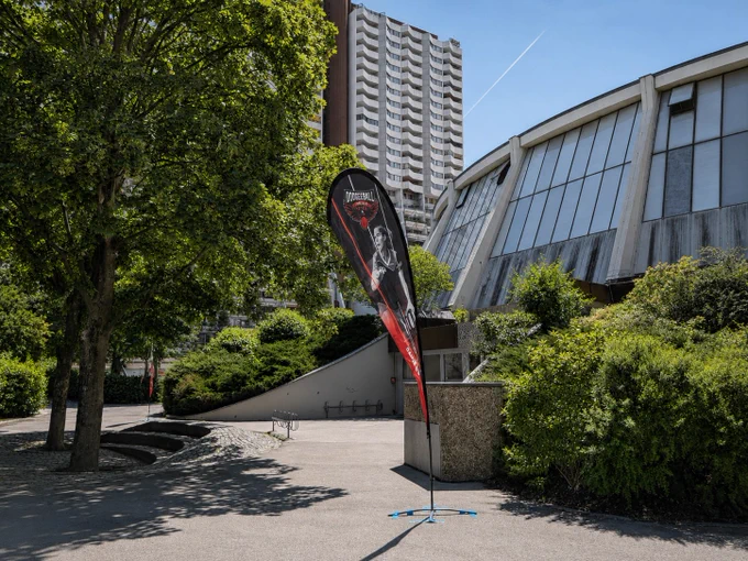 Ein Beachflag mit Dodgeball-Logo steht auf einem Vorplatz. Im Hintergrund ein markantes Gebäude mit geschwungener Glasfassade sowie ein Hochhaus. Grüne Bäume, Sonnenschein.