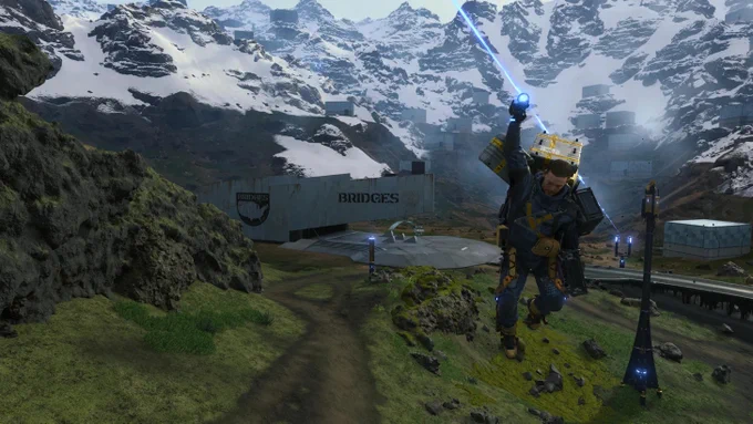 Death Stranding Director's Cut: Sam Porter Bridges mit Frachtausrüstung streckt den Arm aus, blauer Energiestrahl schießt in den Himmel. Bridges-Station und Bergpanorama im Hintergrund. Screenshot.