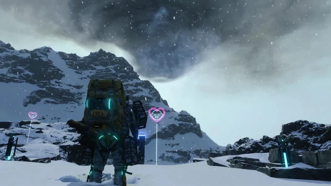 Death Stranding Director's Cut: Sam Porter Bridges mit Exoskelett und schwerem Rucksack im Schneesturm vor Bergkulisse. Pinke Like-Schilder anderer Spieler leuchten im Schnee. Gameplay-Screenshot.