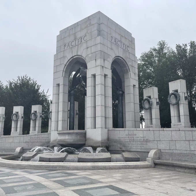 A gray stone tower etched with the word "Pacific" stands at one end of an open stone plaza in an outdoor military memorial.