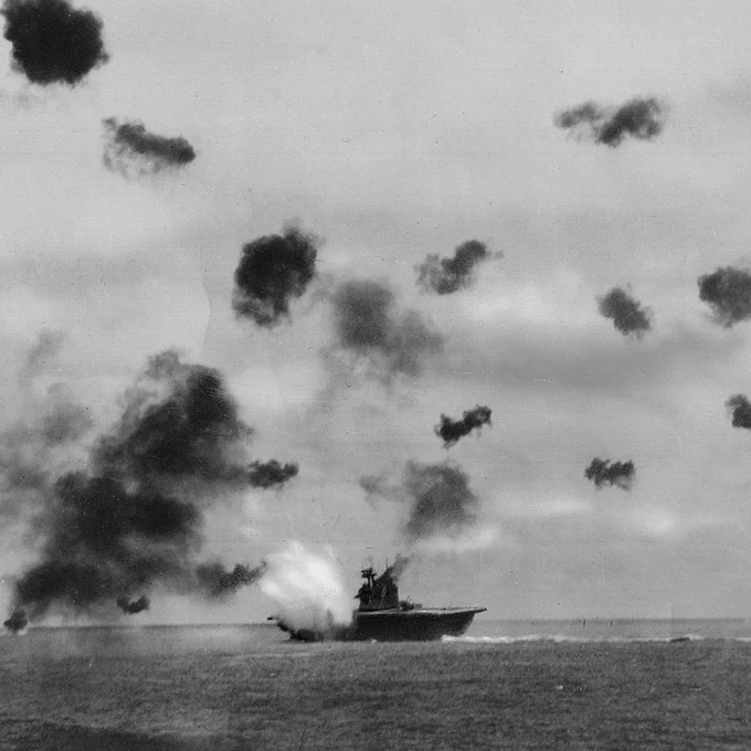 Historic black and white photo of a naval ship under heavy aerial bombardment with smoke bursts surrounding it over the ocean.