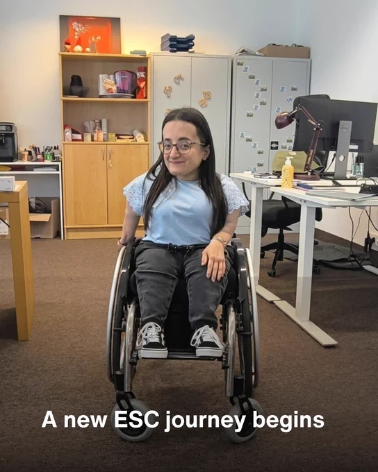 Alt text:

A young woman using a manual wheelchair smiles in an office space. She is wearing glasses, a light blue top, dark jeans and trainers. Office desks, computer monitors, cabinets and shelves are visible in the background. White text at the bottom reads: “A new ESC journey begins.”