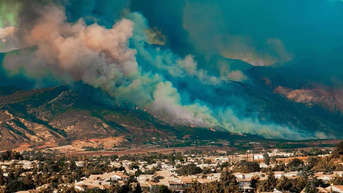 A large plume of smoke billows from a wildfire on a mountainside overlooking a residential area.