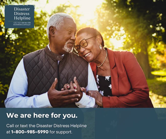 A person with white hair and a beard and another person with short hair are embracing and smiling at each other outdoors during sunset.
