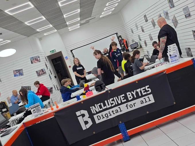 Multiple people behind a desk that says "Inclusive Bytes Digital For All" multiple are on computers, some are looking to be teaching others. Someone behind them all is screaming.