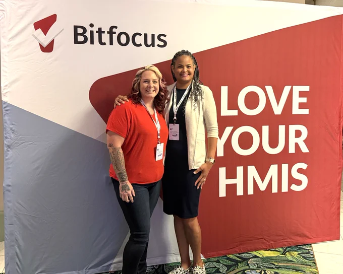 Two women standing in front of a Bitfocus banner that reads 'LOVE YOUR HMIS' at a conference event.