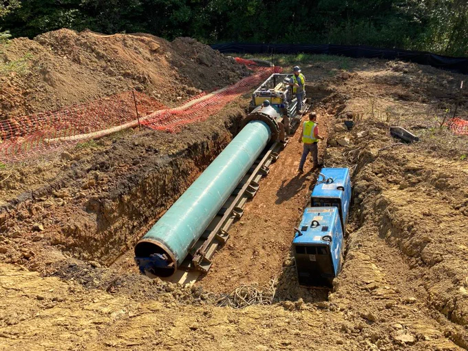 A construction site with workers installing a large green pipeline in a trench. Two blue generator units are nearby. The area is surrounded by dirt piles and safety fencing.
