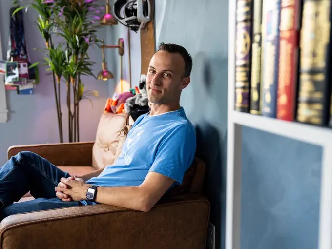 Kent C. Dodds sits on a brown leather couch in his office, leaning back against a dark wall and looking at the camera. A plant, bookshelves, and soft studio lighting frame the relaxed workspace.