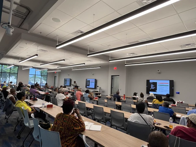 A diverse group of adults attentively watching a presentation on multiple screens in a modern classroom setting.