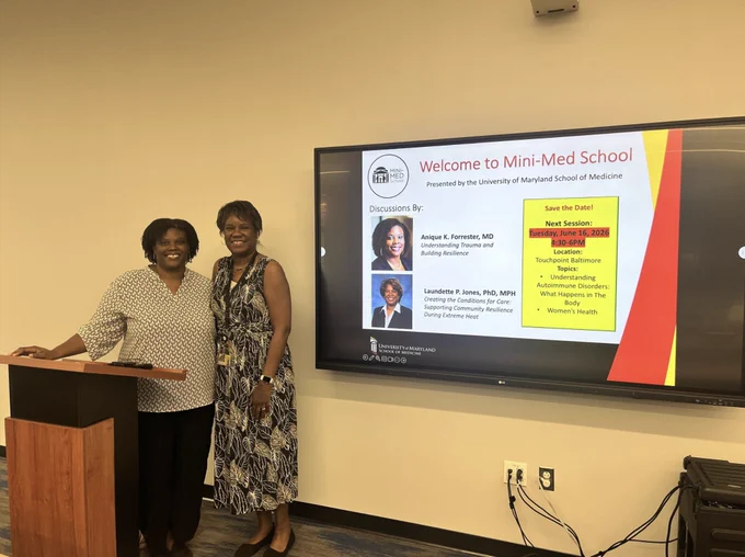 Two women standing next to a podium in a classroom with a screen displaying Mini-Med School session details from University of Maryland School of Medicine.