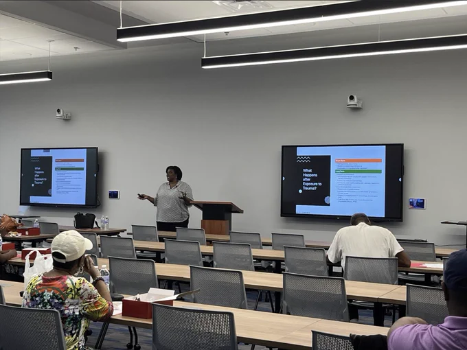Presenter explaining health effects of exposure to trauma in a modern classroom with two large screens displaying slides.